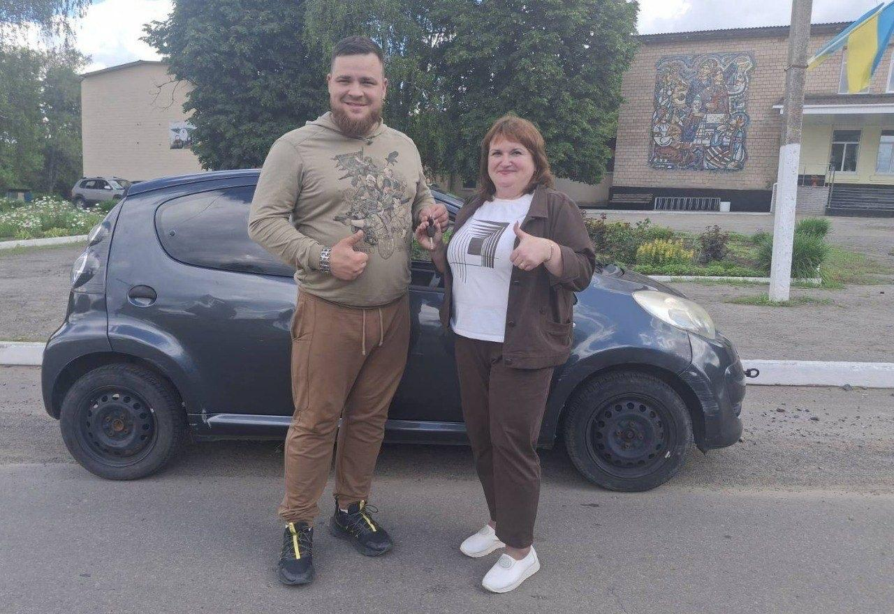Чоловік та жінка стоять поруч із невеликим темно-сірим автомобілем, показуючи жест «великий палець угору», при цьому жінка тримає в руці ключі від авто.