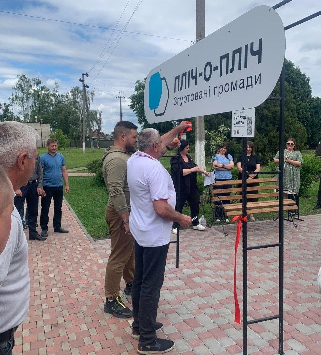 Група людей зібралася на відкритому повітрі біля великої білої вивіски з написом «ПЛІЧ-О-ПЛІЧ згуртовані громади» під час урочистого перерізання червоної стрічки.