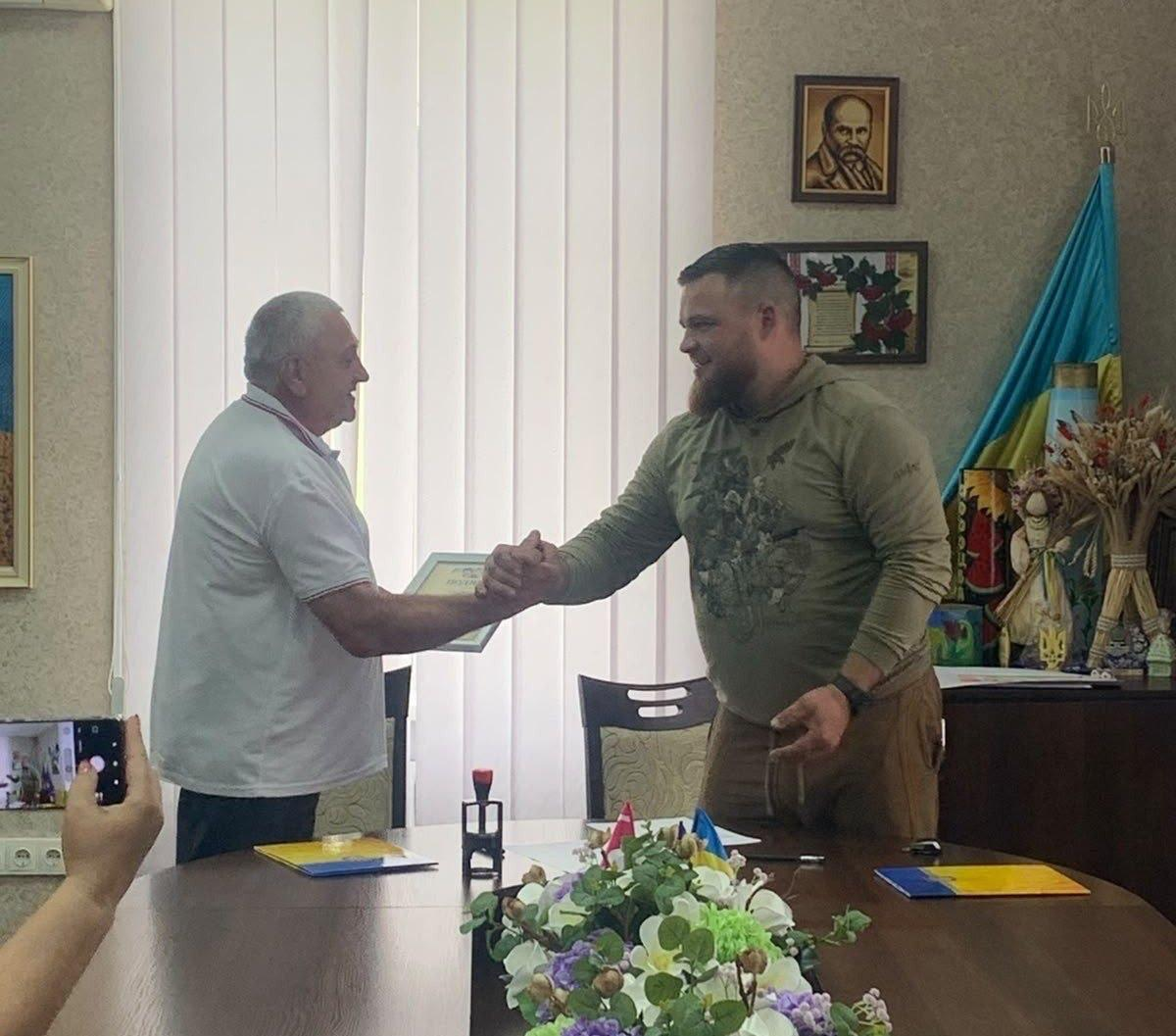 Двоє чоловіків тиснуть один одному руки над робочим столом у кабінеті, при цьому один із них передає іншому грамоту або диплом у рамці.