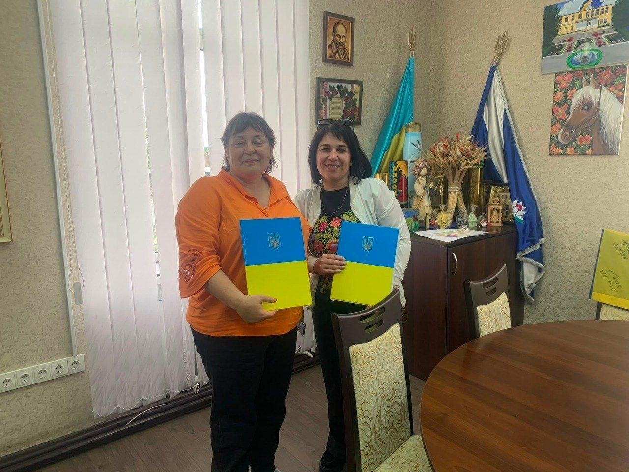 Дві жінки стоять у робочому кабінеті, тримаючи в руках синьо-жовті папки з гербом України, на тлі прапорів, картини та портрета на стіні.