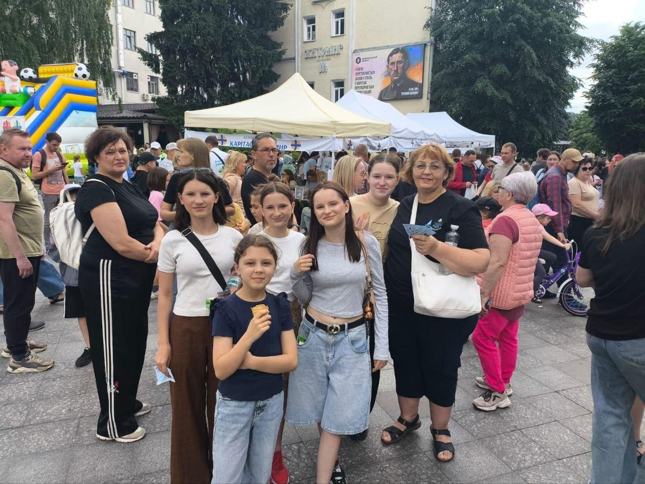Група жінок та дітей позирует для фото на багатолюдній міській площі під час масового заходу з білими наметами та надувним батутом на задньому плані.