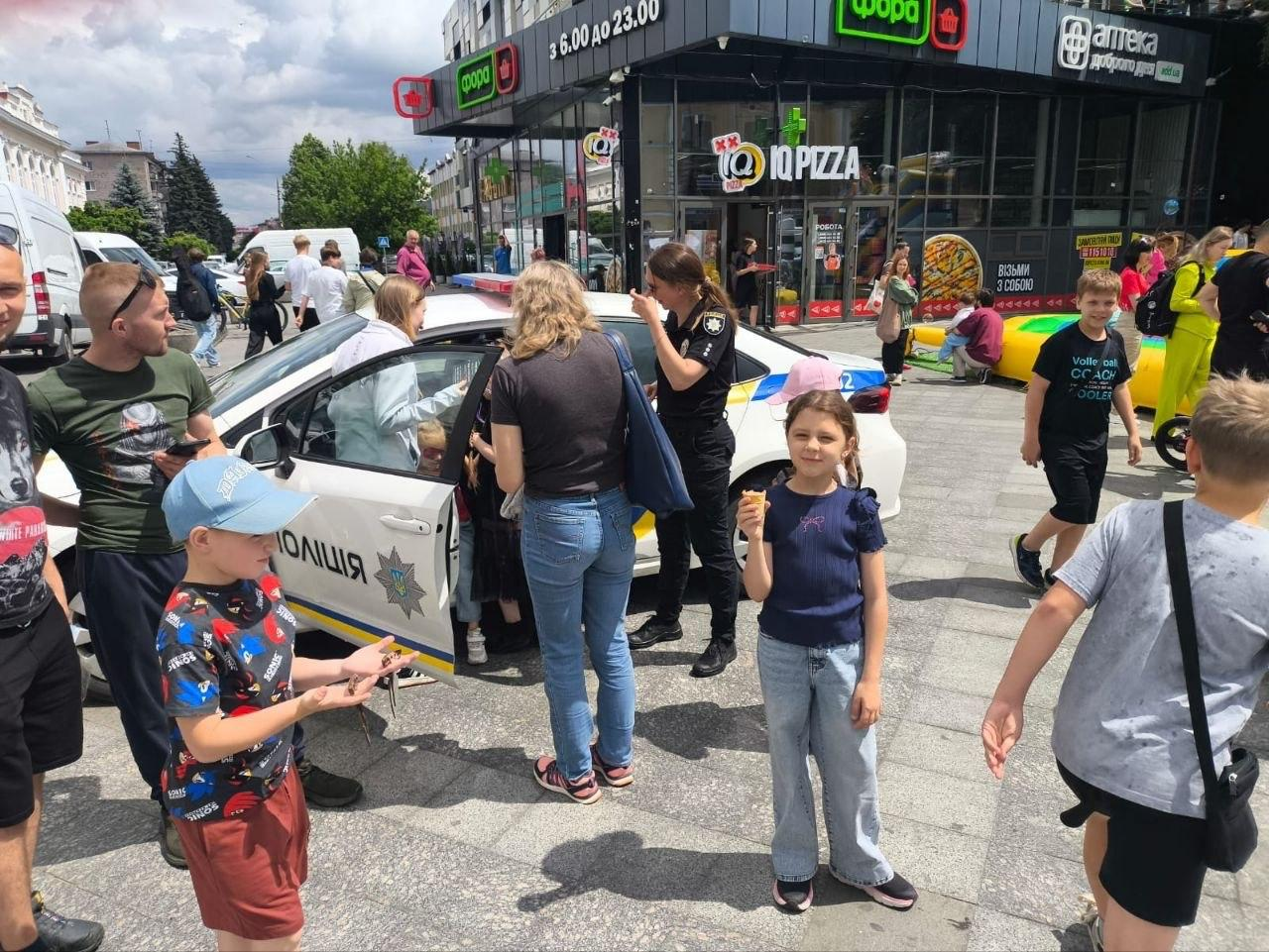 Діти та дорослі зібралися біля білого патрульного автомобіля поліції на міській площі, поруч із працівницею поліції у формі, на тлі будівлі з вивісками торговельних закладів.