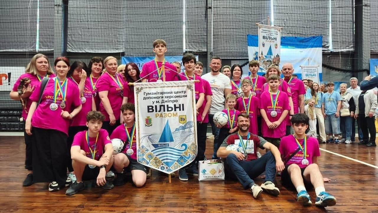 Велика група дітей та дорослих у яскраво-рожевих спортивних футболках із медалями позує для спільного фото у спортивному залі навколо великого декоративного банера з написом «ВІЛЬНІ РАЗОМ».