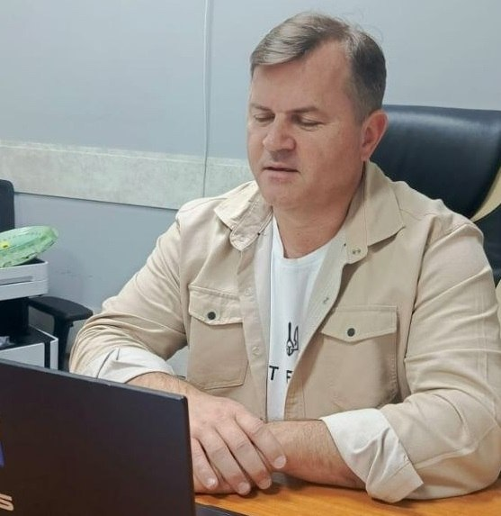 Чоловік у бежевій сорочці поверх білої футболки сидить за дерев'яним столом перед відкритим ноутбуком у робочому кабінеті.