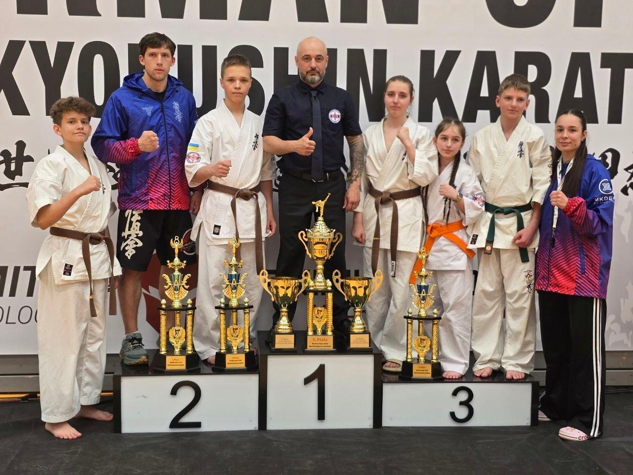 Група спортсменів-каратистів у білих кімоно та тренерів стоїть на п'єдесталі пошани з великими золотими кубками на тлі банера з написом «KYOKUSHIN KARATE».