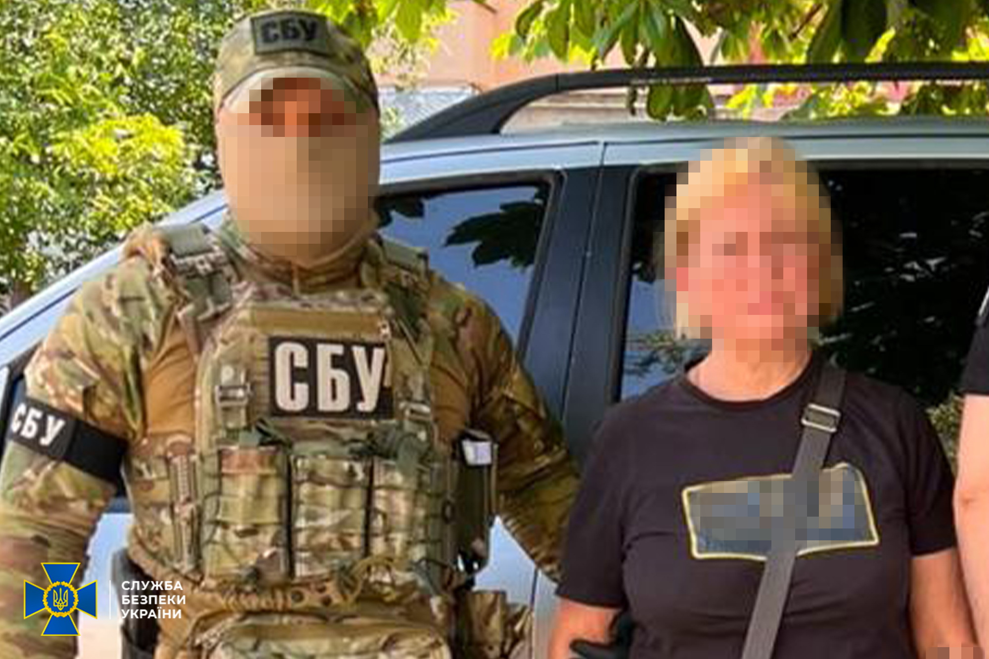 Військовослужбовець Служби безпеки України у камуфляжній формі та балаклаві стоїть поруч із жінкою на тлі автомобіля; обличчя обох осіб на фотографії повністю розмиті та приховані.
