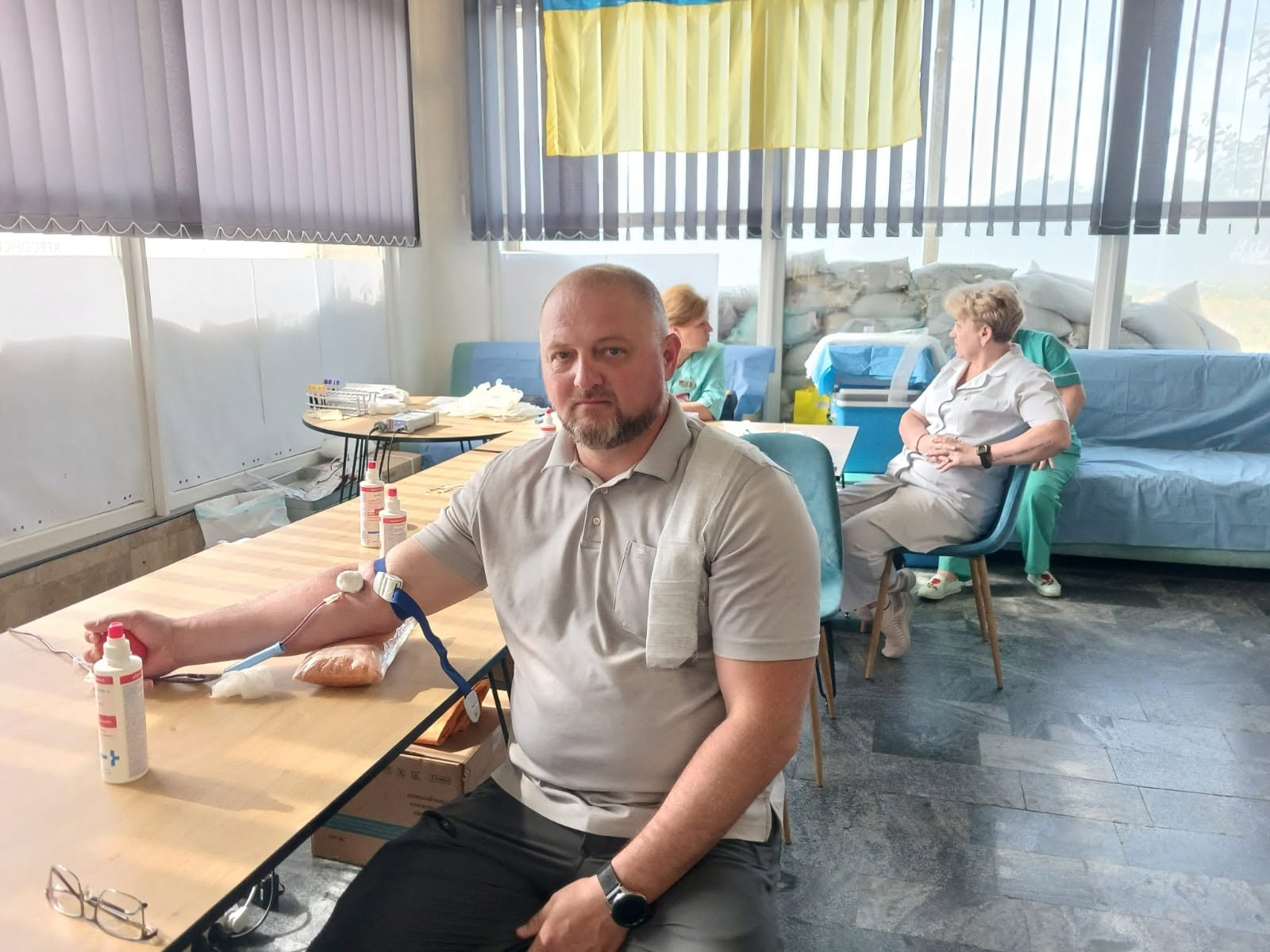 Чоловік у сірій футболці-поло сидить за столом у приміщенні з прапором України та здає кров, поки на задньому плані медичні працівники спілкуються з іншим донором.