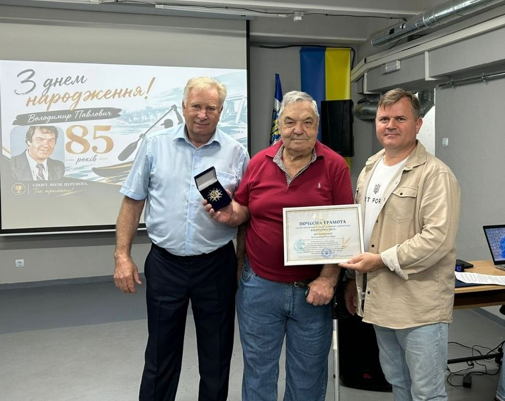 Три чоловіки стоять у приміщенні на тлі великого екрана з вітальним написом «З днем народження! Володимир Павлович 85 років»; чоловік по центру тримає почесну грамоту, а чоловік ліворуч демонструє відкриту коробочку з нагородою.