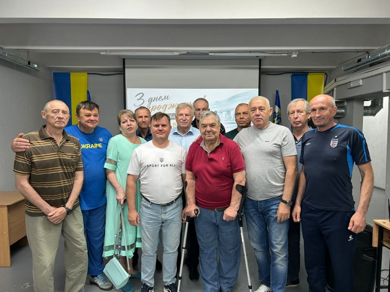 Велика група людей позує для спільного фото в приміщенні на тлі екрана з вітальним написом «З днем народження!», де по центру стоїть чоловік похилого віку з двома підліктьовими милицями, а обабіч розміщено прапори України.