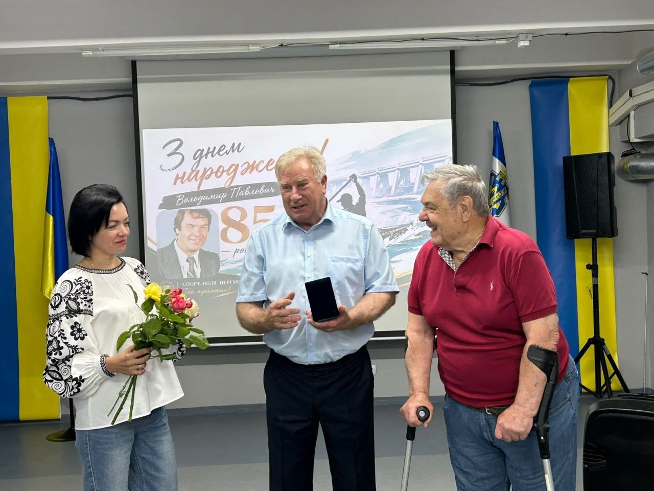 Жінка у вишиванці з букетом квітів та двоє чоловіків спілкуються в приміщенні на тлі екрана з написом «З днем народження! Володимир Павлович 85 років», при цьому чоловік по центру тримає коробочку з нагородою, а чоловік праворуч спирається на милиці.