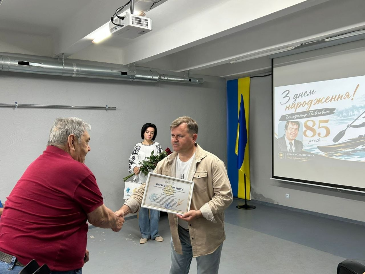Чоловік у світлій сорочці тисне руку чоловікові в бордовій футболці та вручає йому почесну грамоту на тлі великого екрана з вітальним написом «З днем народження! Володимир Павлович 85 років», поки на задньому плані стоїть жінка у вишиванці з букетом квітів.