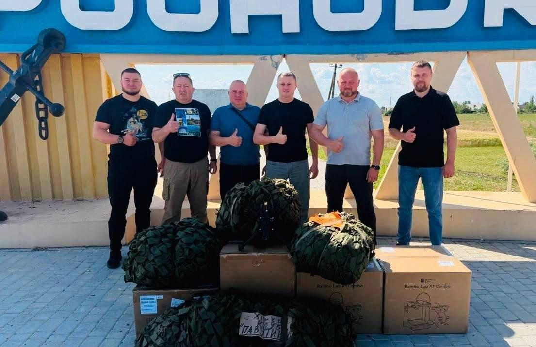 Шість чоловіків стоять у ряд на вулиці біля великого синьо-жовтого знака, показуючи жест «великий палець угору», перед ними на землі складені картонні коробки з написом «Bambu Lab» та військове спорядження у маскувальних сітках.