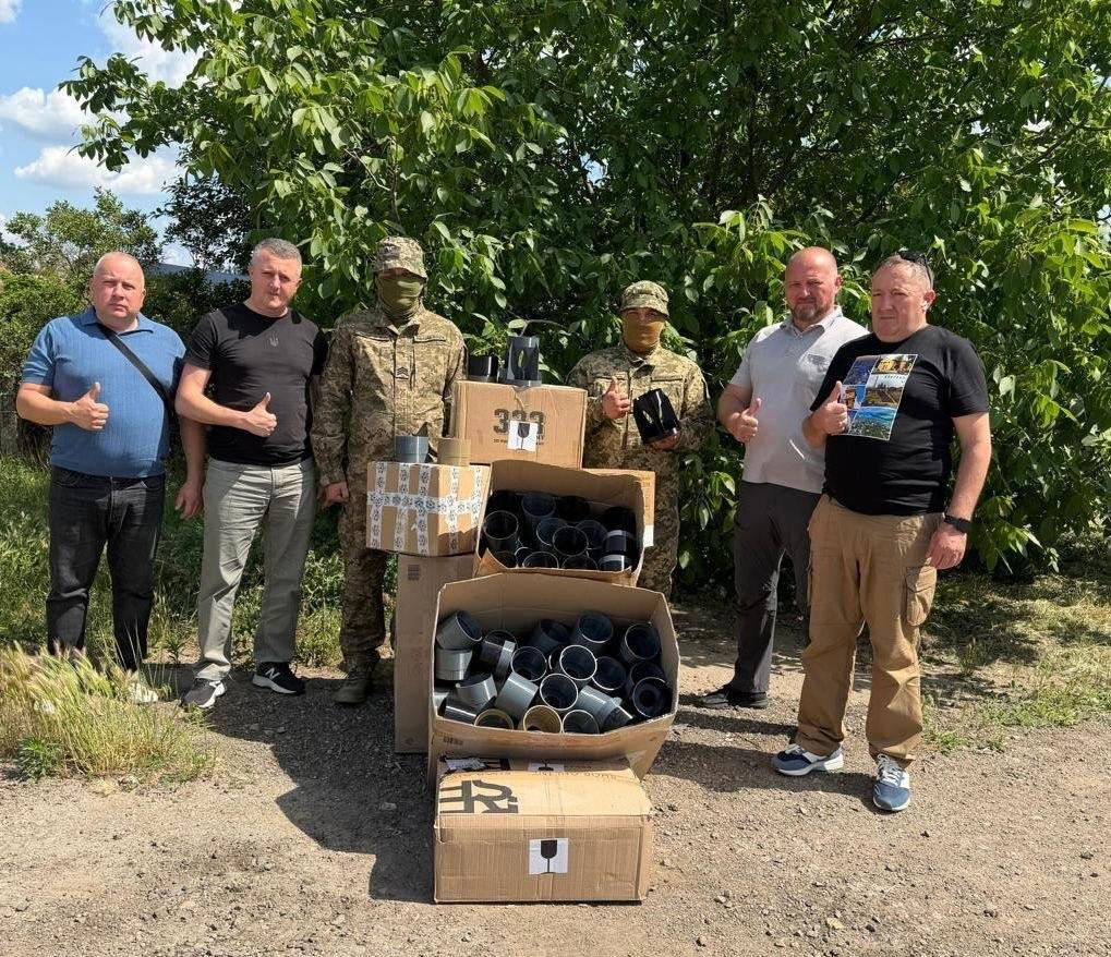 Група з чотирьох цивільних чоловіків та двох військовослужбовців у камуфляжі й балаклавах позує на тлі зелених дерев поруч із великими картонними коробками, що заповнені циліндричними пластиковими деталями.