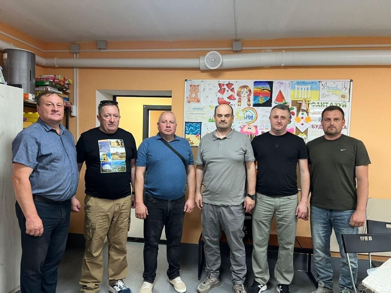 Шість чоловіків у повсякденному одязі стоять у ряд у кімнаті на тлі стіни, на якій розміщено великий стенд із дитячими малюнками та аплікаціями.