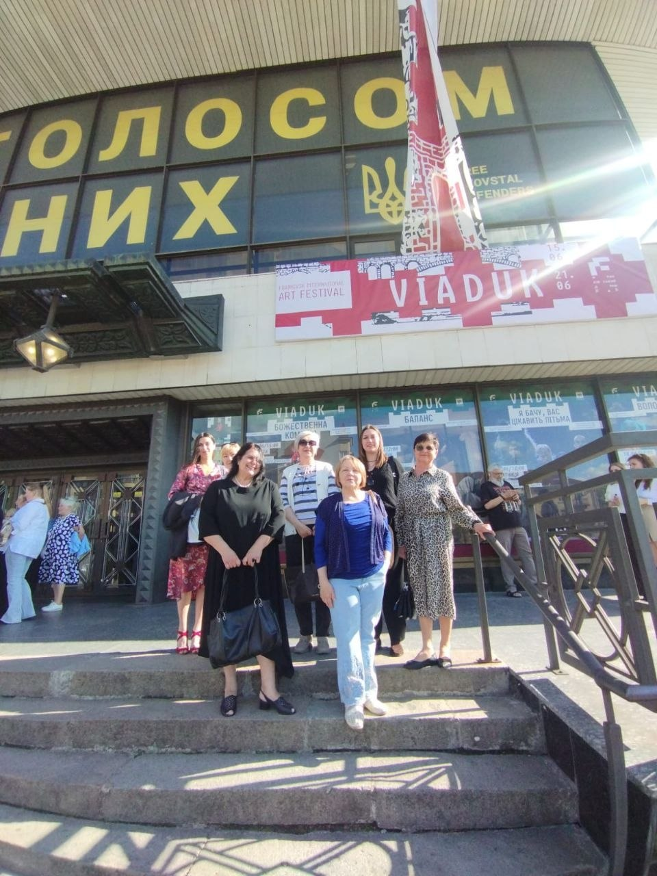 Група жінок позує на кам'яних сходах перед входом до будівлі кінотеатру імені Івана Миколайчука з великими написами «VIADUK» та «ART FESTIVAL» на фасаді й вікнах.