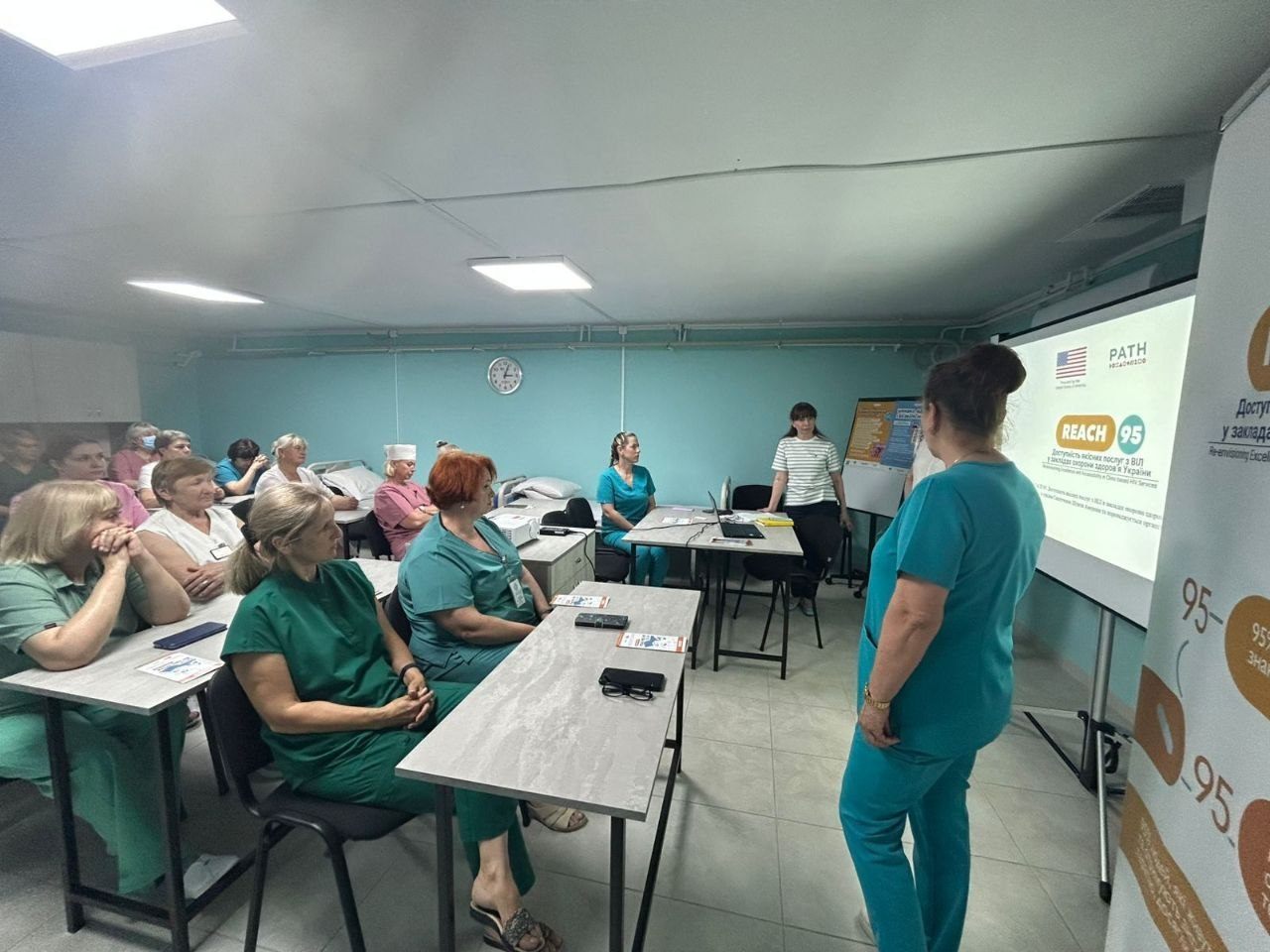 Медичні працівники слухають лекцію в навчальній аудиторії, сидячи за партами перед проєкційним екраном з логотипами «PATH» та «REACH 95», біля якого стоїть спікерка в бірюзовому медичному костюмі.