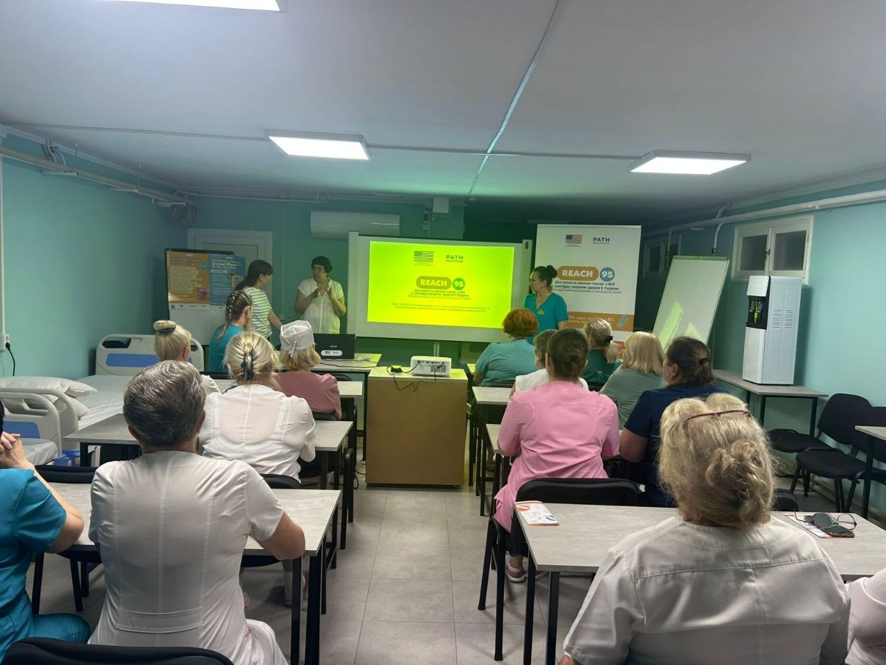 Презентація для медичних працівників у навчальному кабінеті, де слухачі сидять за партами спиною до камери, а попереду розміщено яскраво-жовтий проєкційний екран та банер з логотипами «PATH» та «REACH 95».