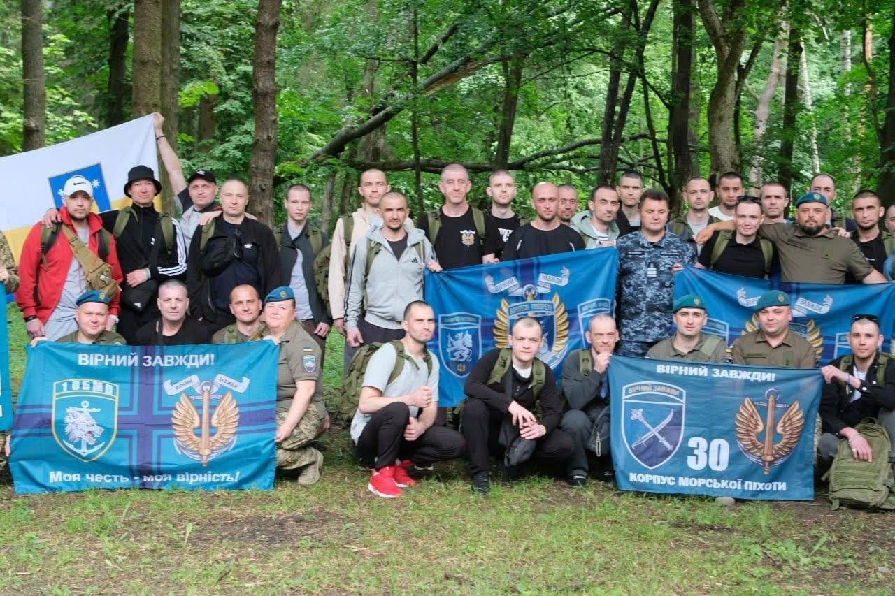 Велика група чоловіків у військовому та цивільному одязі позує для спільного фото в лісі, тримаючи прапори з символікою Морської піхоти України, емблемами підрозділів та написами «ВІРНИЙ ЗАВЖДИ!».