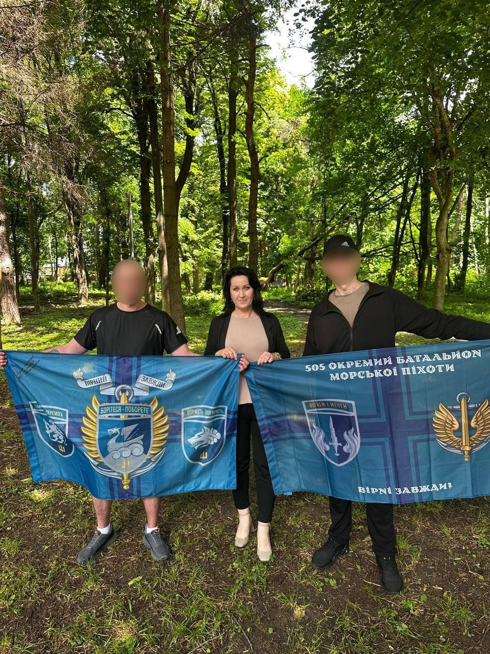 Жінка та двоє чоловіків із розмитими обличчями стоять у парку й тримають два синьо-зелені прапори з символікою 505-го окремого батальйону морської піхоти та написами «БОРІТЕСЯ – ПОБОРЕТЕ» і «ВІРНІ ЗАВЖДИ!».