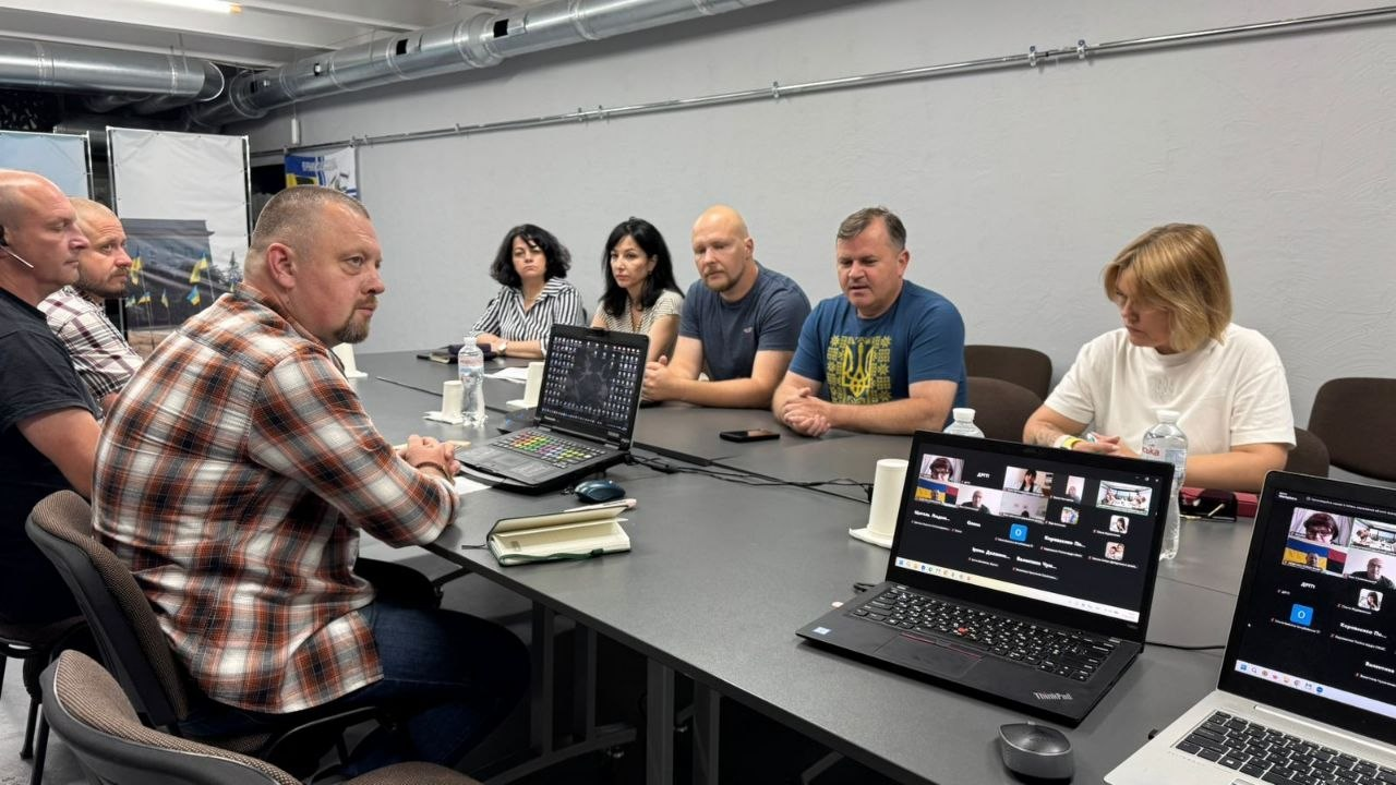 Група людей сидить навколо великого столу в робочому кабінеті під час наради, де на столі розставлені відкриті ноутбуки, один з яких показує вікна онлайн-дзвінка у Zoom.