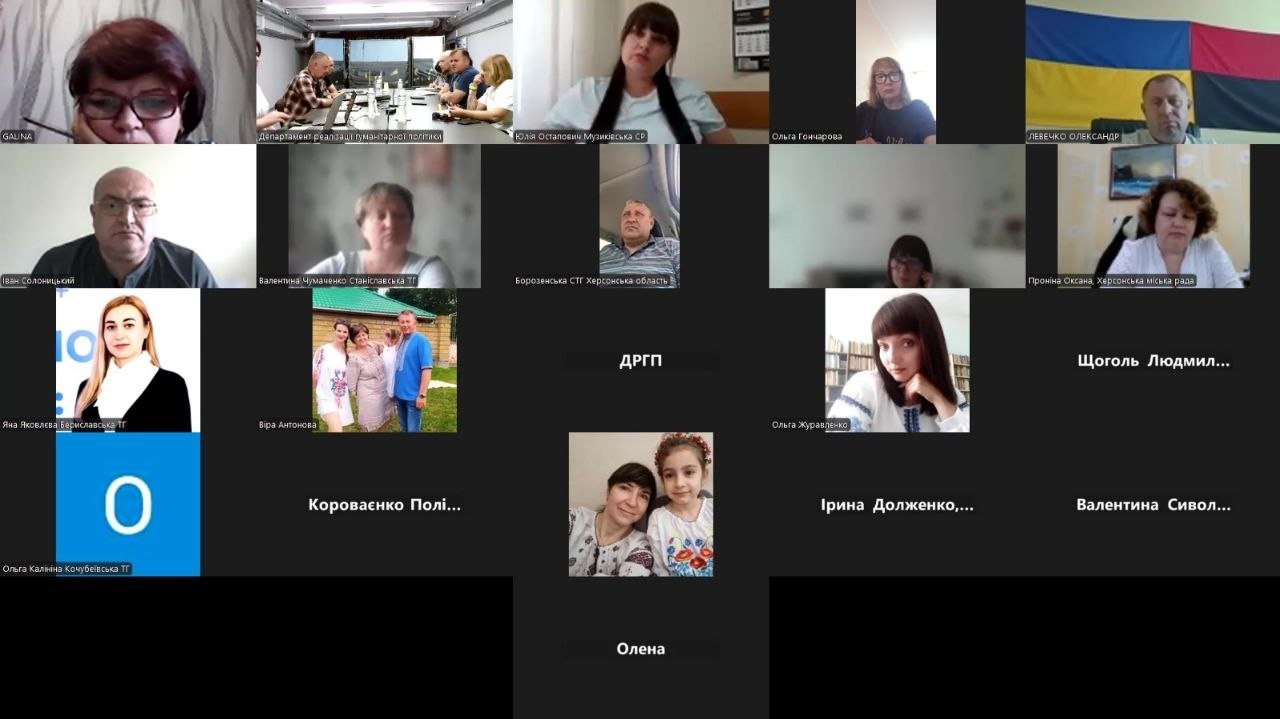 Скриншот онлайн-наради в Zoom із плитковим інтерфейсом, де відображаються віконця учасників з відео та фотографіями профілів, зокрема деякі люди у вишиванках, а в одному з вікон видно кабінет з прапорами України.