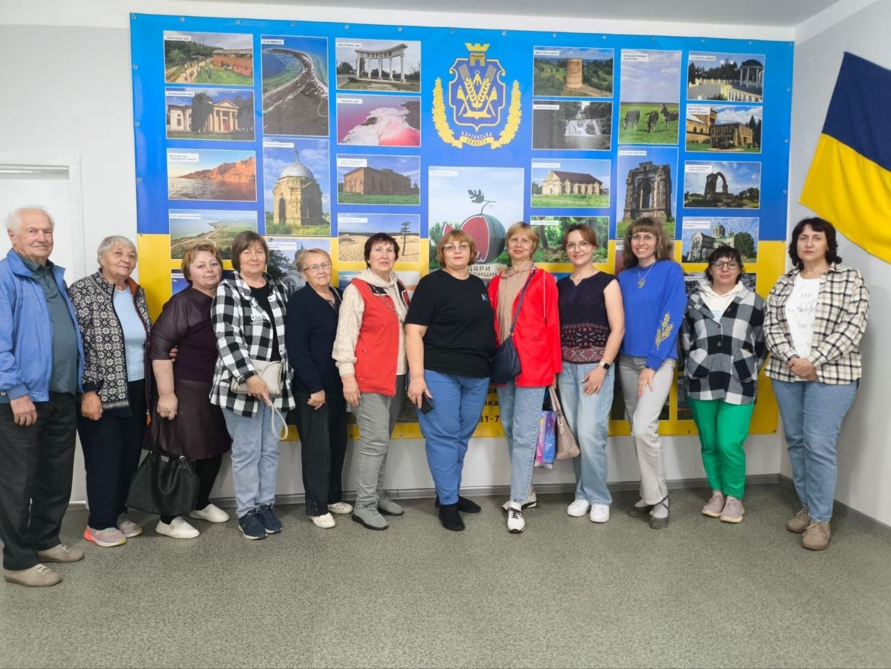 Група людей похилого та середнього віку стоїть у ряд і позує для фотографії на тлі великого жовто-блакитного патріотичного стенда з фотографіями пам'яток та гербом.