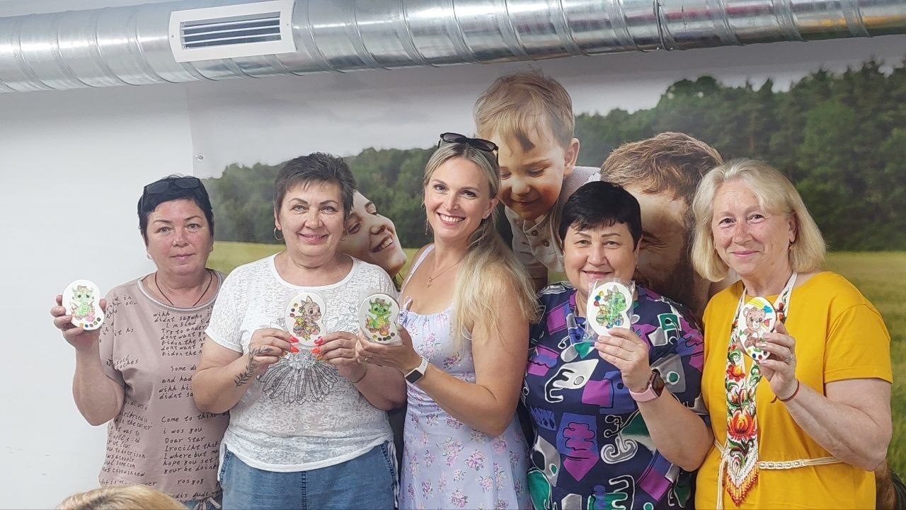 Група з п'яти жінок посміхається і позиє для фотографії, тримаючи в руках розписані декоративні пряники або сувеніри, на тлі великих фотошпалер із зображенням природи та людей.