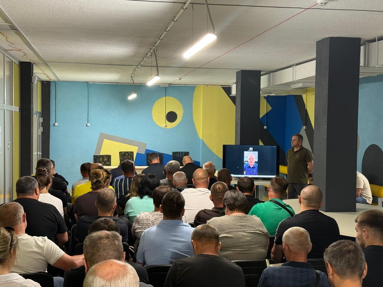 Група людей сидить спиною до камери в залі з яскраво розписаними стінами та слухає виступ чоловіка в мілітарі-одязі, поруч із яким розташовано великий екран із відеозв'язком онлайн-спікера.