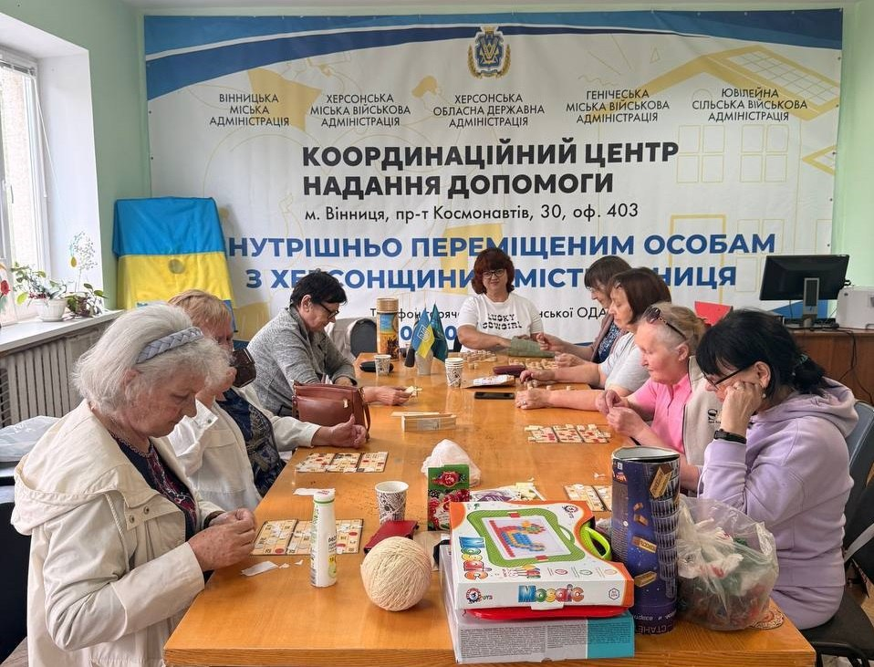Група жінок сидить за довгим столом і грає в настільні ігри (лото) у приміщенні Координаційного центру надання допомоги внутрішньо переміщеним особам з Херсонщини.