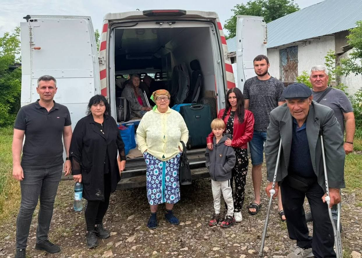 Група людей різного віку, серед яких є літні громадяни, дитина та чоловік на милицях, позує для фото біля білого мікроавтобуса з відкритими задніми дверима, завантаженого валізами та речами.