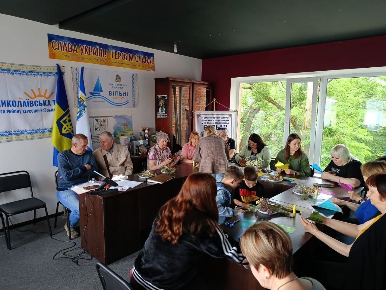 Група людей різного віку, серед яких дорослі та діти, сидить за великим столом у світлій кімнаті з патріотичними банерами та займається творчістю, виготовляючи вироби з паперу та квітів.