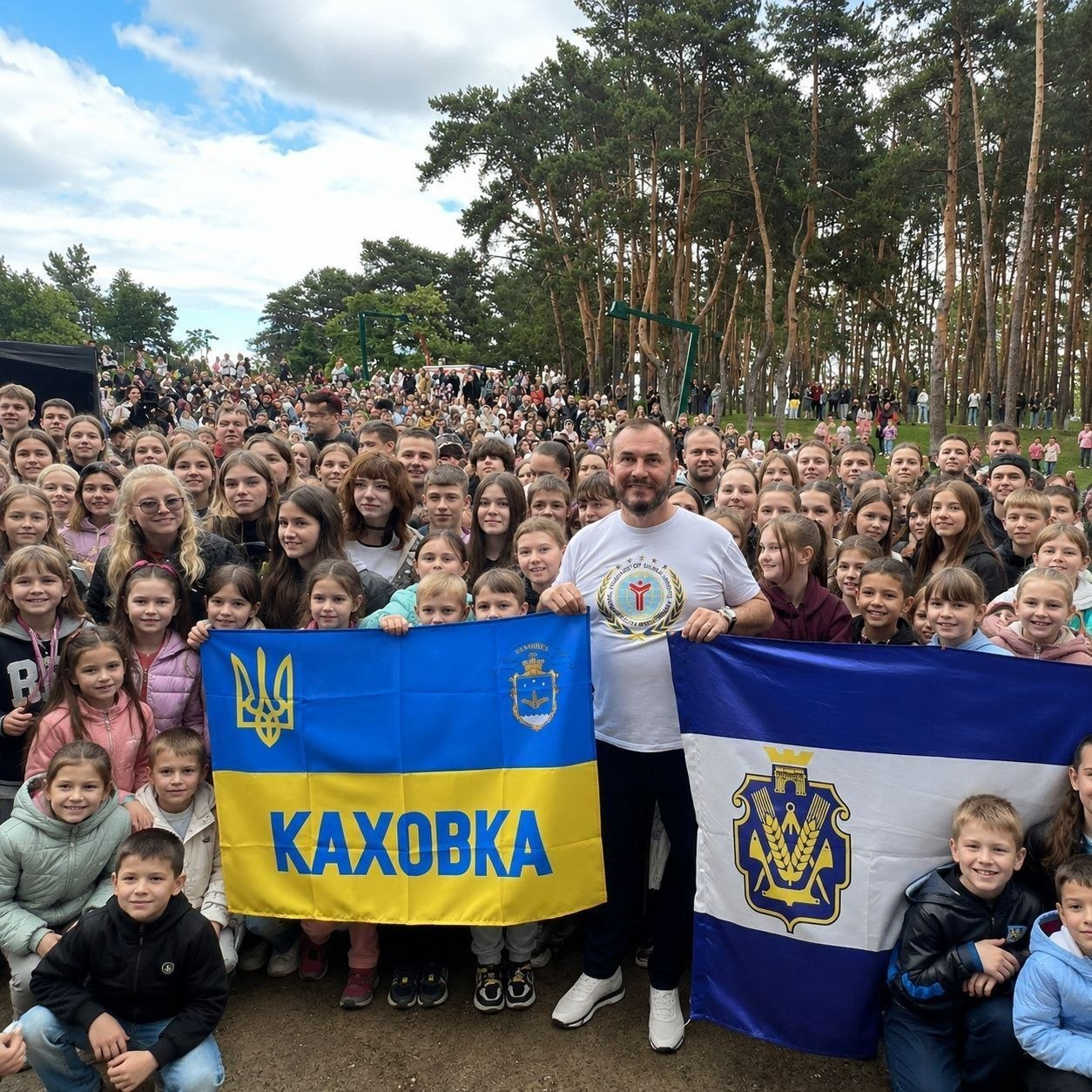 Група дітей та дорослих на природі тримає синьо-жовтий прапор із написом «КАХОВКА» та синьо-білий прапор із гербом міста.