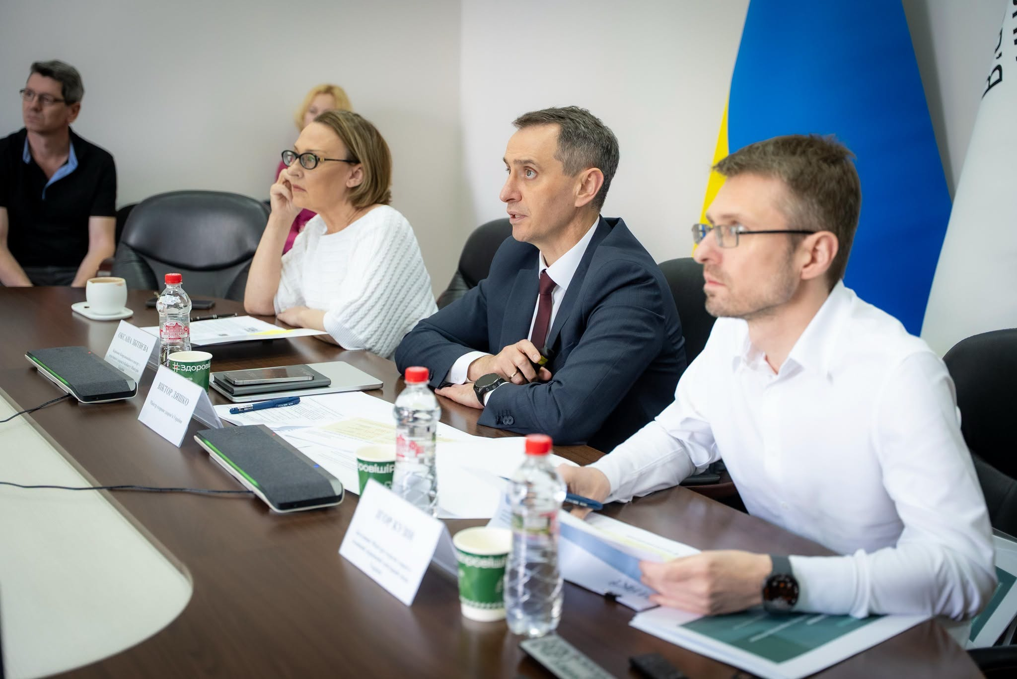 Ділова зустріч у кабінеті, де за великим столом сидять та обговорюють робочі питання чоловіки та жінки в офіційному вбранні на тлі прапора України.