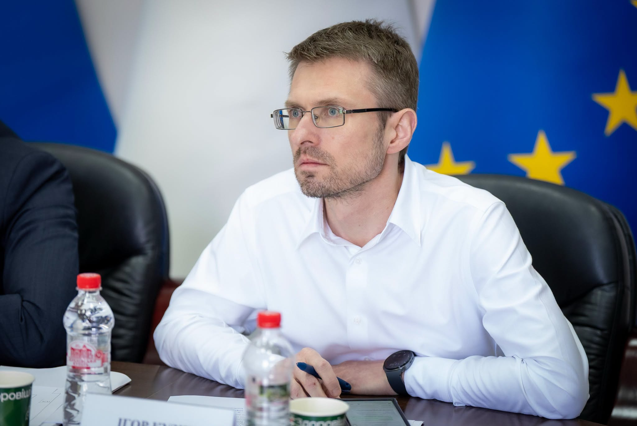 Чоловік в окулярах та білій сорочці сидить за столом під час офіційного заходу на тлі прапора Європейського Союзу.