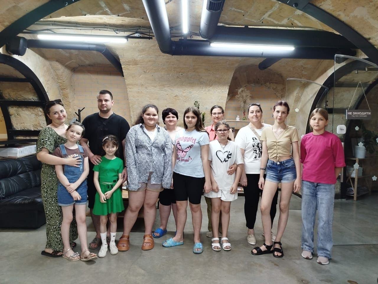 Група дорослих та дітей стоїть разом для спільного фото у приміщенні з арочною стелею, що нагадує облаштоване укриття.
