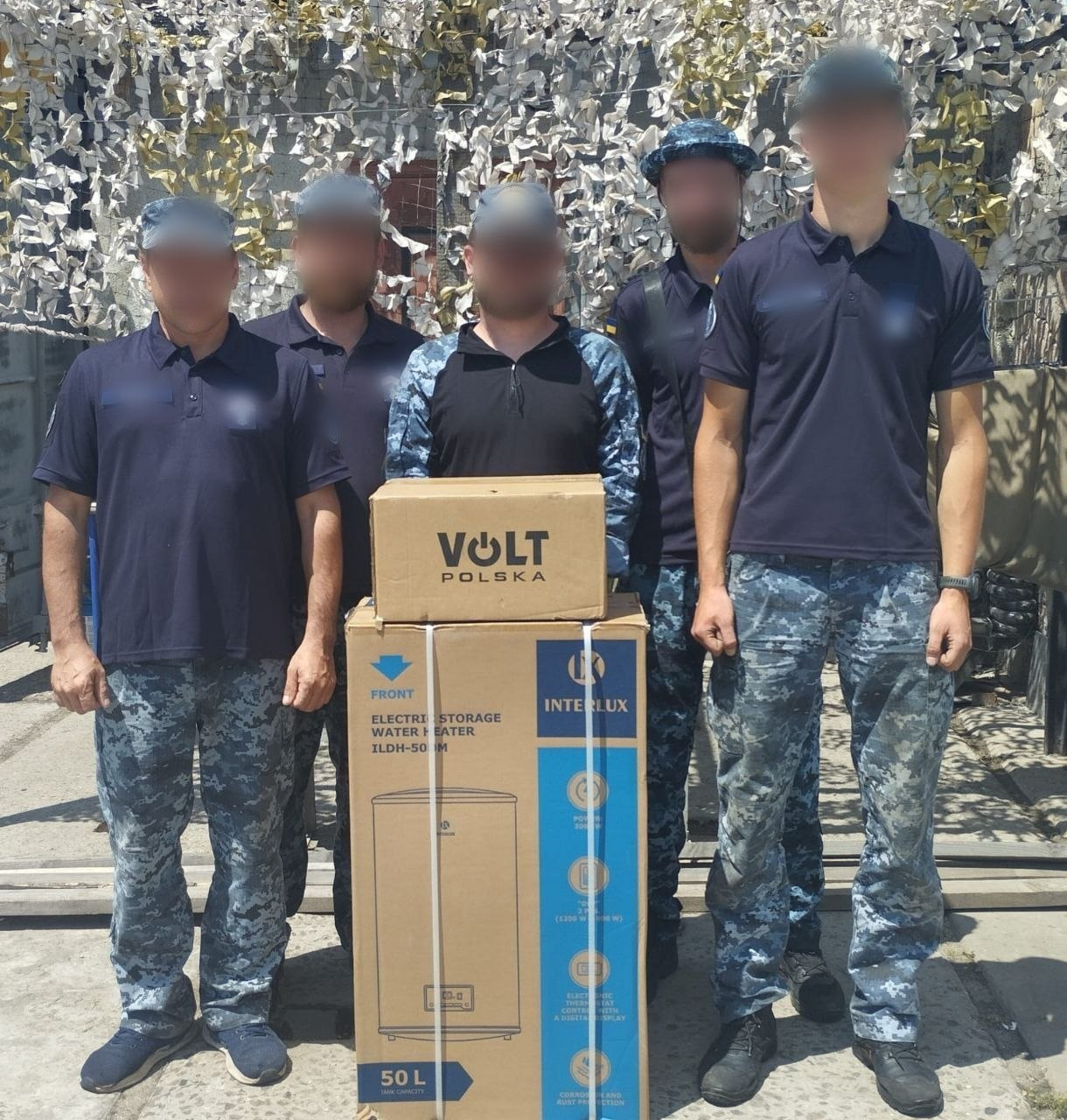 П'ятеро чоловіків у військовій або службовій формі з камуфляжними штанами стоять на вулиці перед маскувальною сіткою. Попереду них розташовані дві великі картонні коробки: нижня — з малюнком бойлера бренду «INTERLUX» на 50 літрів, верхня — з логотипом «VOLT POLSKA».