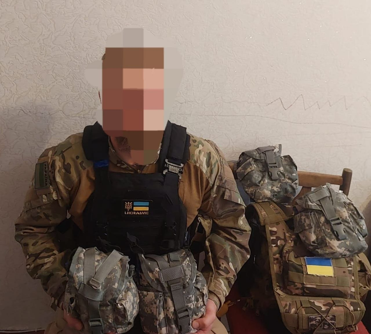 Чоловік у камуфляжній формі та чорній тактичній розгрузці з прапором України тримає в руках військове спорядження, сидячи в приміщенні біля стільця з наплічником та захисним шоломом у камуфляжному чохлі.