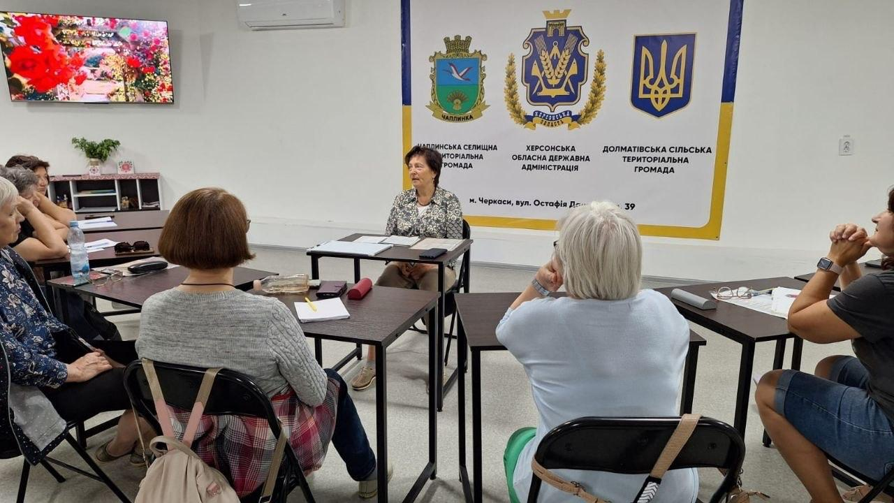 Жінки сидять за столами в офісному приміщенні та слухають доповідачку. На стіні позаду висить банер із гербами громад Херсонської області та телевізор. На столах лежать записники й особисті речі.