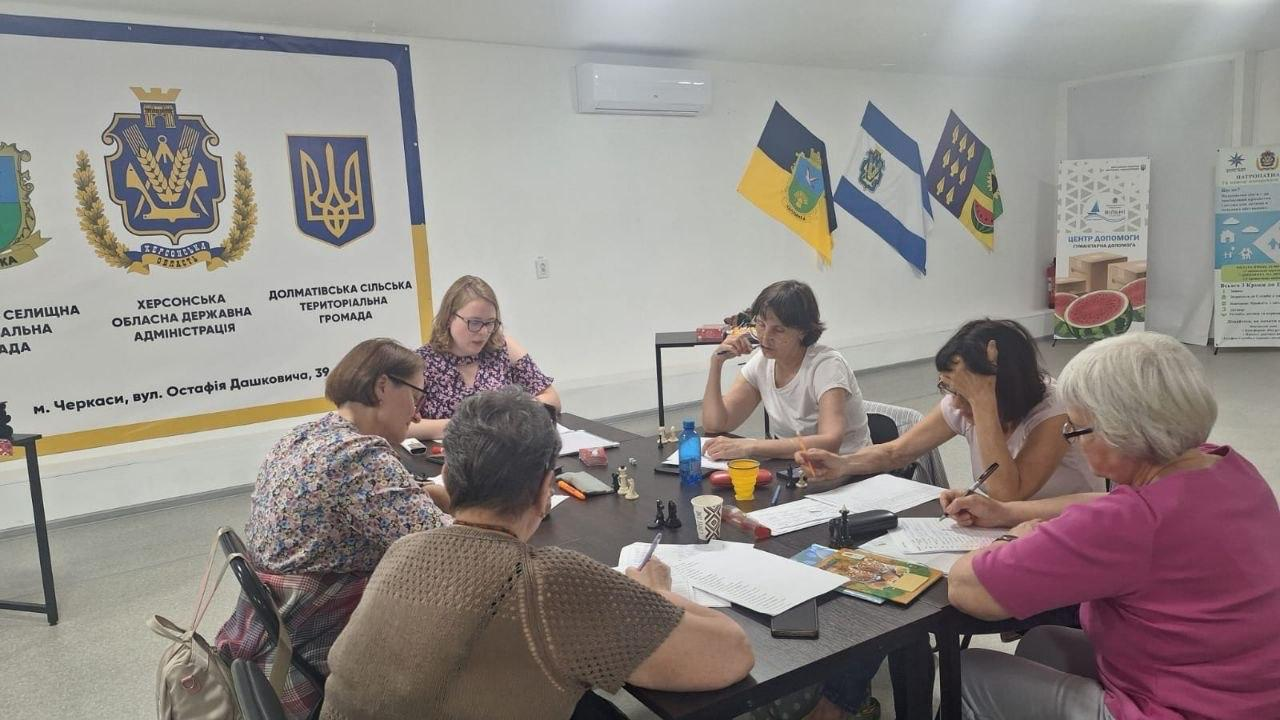 Група жінок сидить навколо великого столу в офісному приміщенні під час зустрічі. На стіні позаду висить банер із гербами Херсонської області та територіальних громад, а поруч закріплені кілька прапорів. На столі розкладені папери, канцелярія, пляшки з водою та шахові фігури.