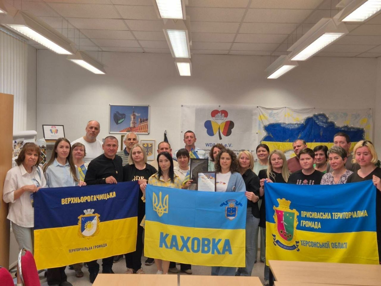 Група людей стоїть у приміщенні та тримає три прапори територіальних громад Херсонської області: Верхньорогачицької, Каховської та Присиваської.