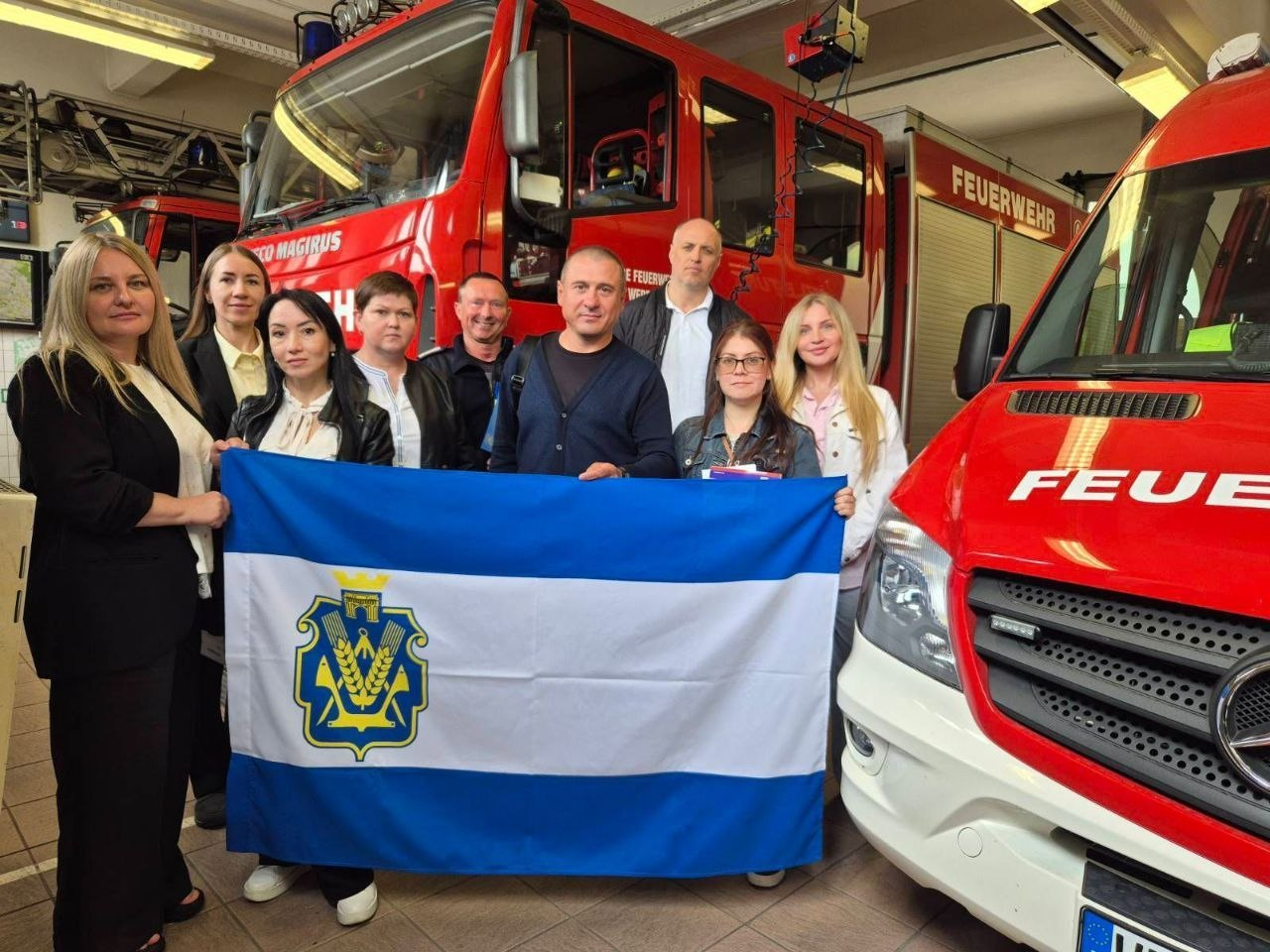 Група людей стоїть у пожежному депо на тлі червоних пожежних автомобілів і тримає синьо-білий прапор із гербом Херсонської області.