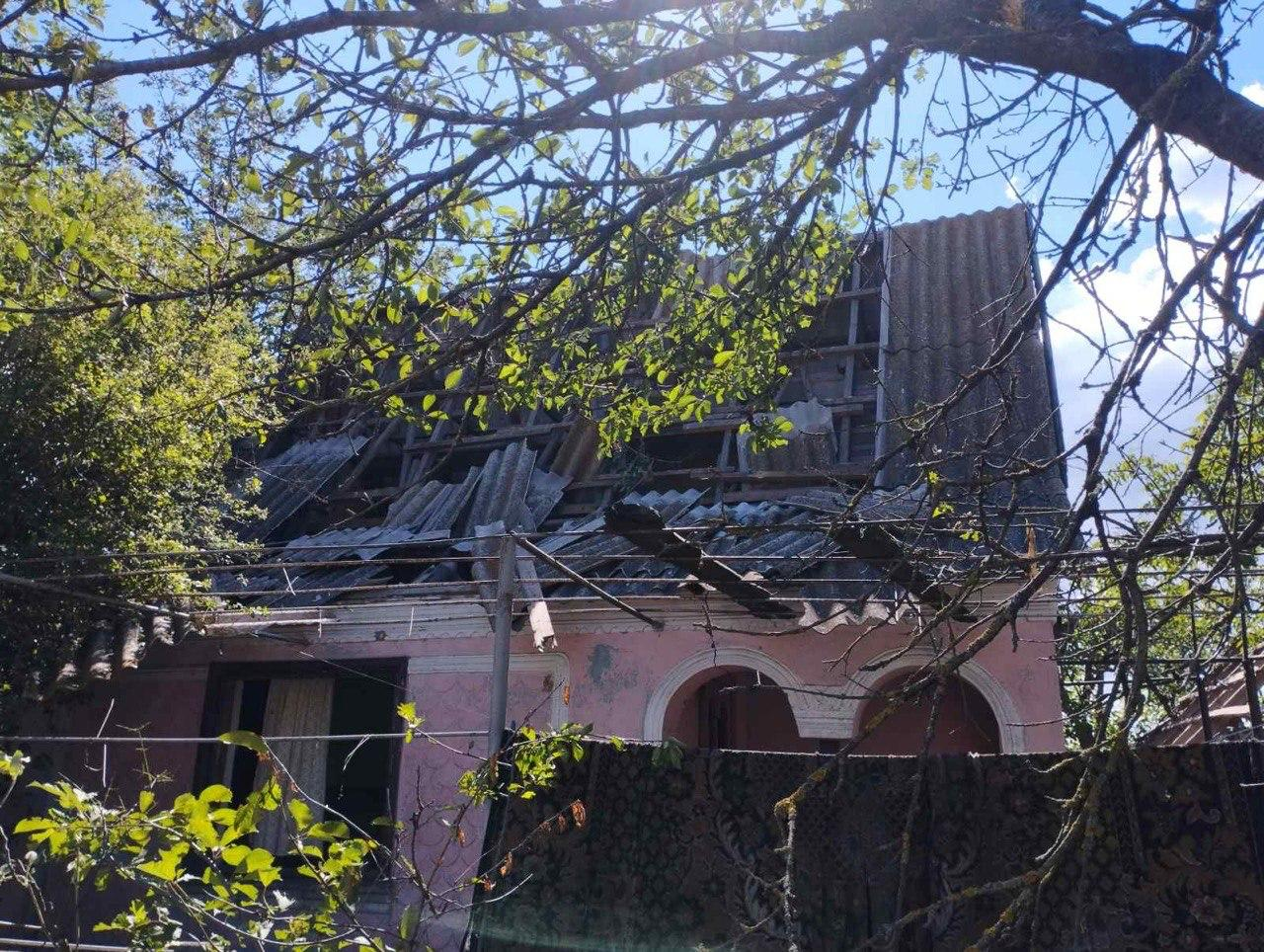Двоповерховий рожевий будинок із сильно зруйнованим шиферним дахом та оголеними дерев'яними балками, знятий крізь гілки зелених дерев.