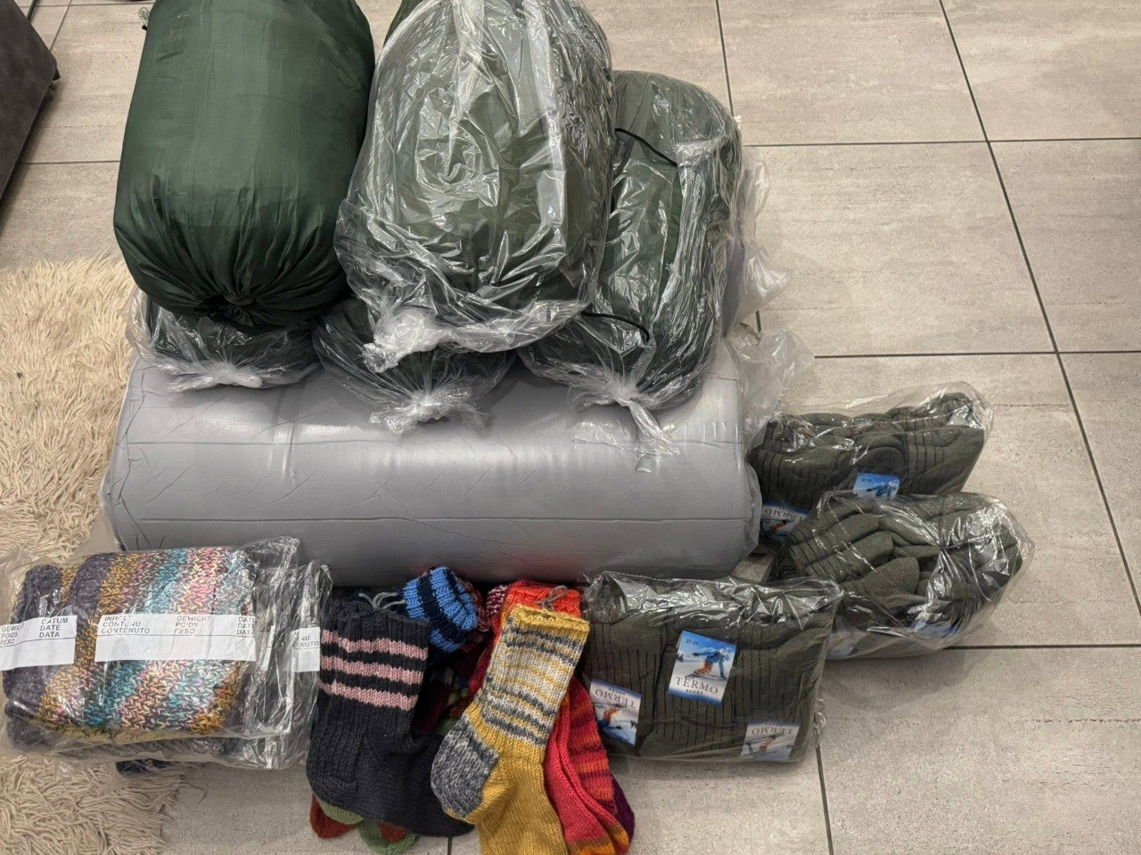 Волонтерська допомога на кахельній підлозі: три зелені спальні мішки у чохлах, великий сірий згорток, в'язані кольорові шкарпетки, а також упаковані термошкарпетки та рукавиці.