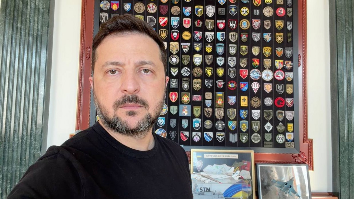 Чоловік у чорній футболці на тлі великого настінного стенда з численними шевронами та військовими патчами. Поруч стоять невеликі рамки із зображеннями літаків та патріотичною символікою.