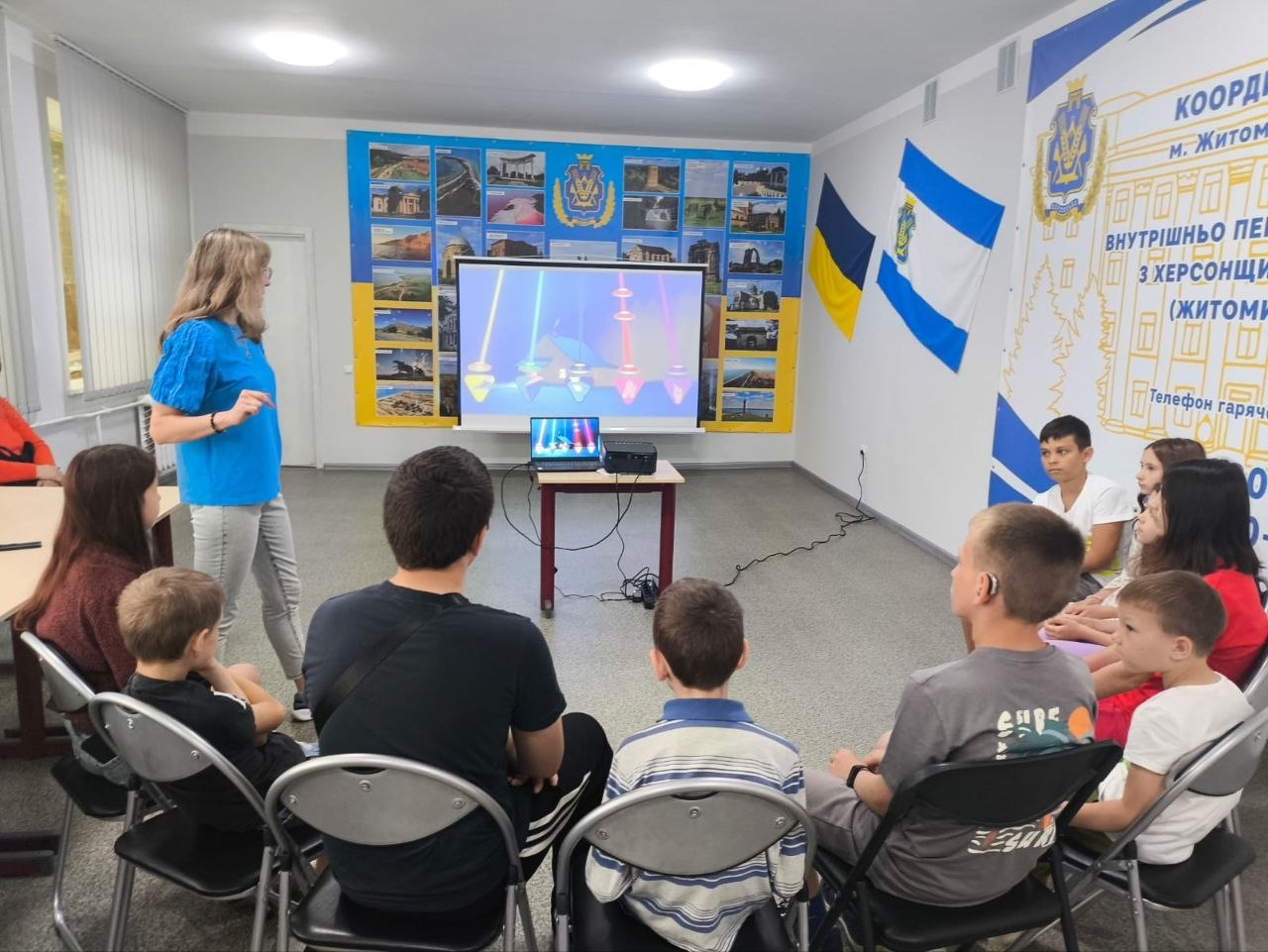 Група дітей та дорослих сидить півколом у світлій кімнаті та дивиться на великий екран, де демонструється яскрава презентація або гра. Зліва стоїть жінка у блакитній блузці, яка веде захід. На стінах кімнати розміщені плакати з фотографіями, інформаційний стенд та прапори України.