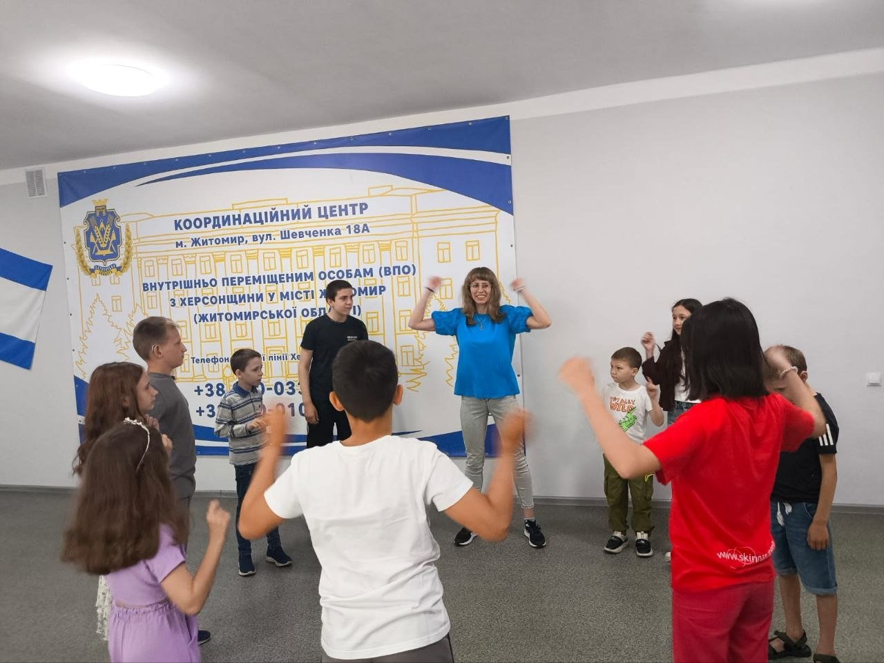 Група дітей та дорослих роблять руханку під час заняття у просторому приміщенні. По центру жінка показує рухи, які повторюють діти навколо неї. На задньому плані розміщено великий інформаційний банер Координаційного центру.