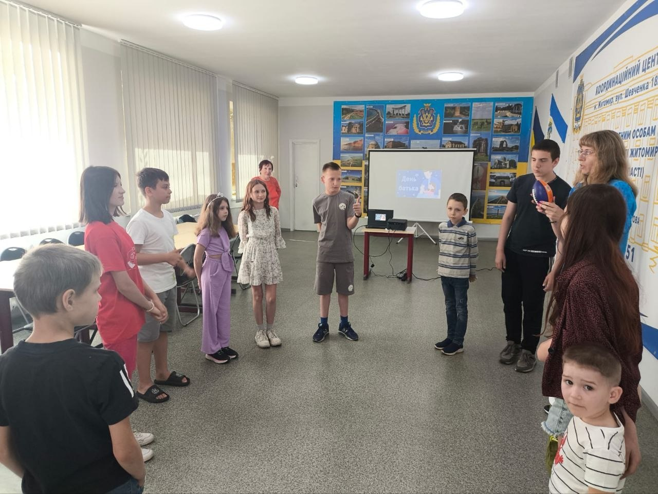 Група дітей та дорослих стоїть півколом у світлій кімнаті під час заходу. У центрі хлопчик тримає мікрофон і виступає, а на задньому плані видно екран із презентацією. На стінах розміщені інформаційні стенди, банер Координаційного центру та прапори України.
