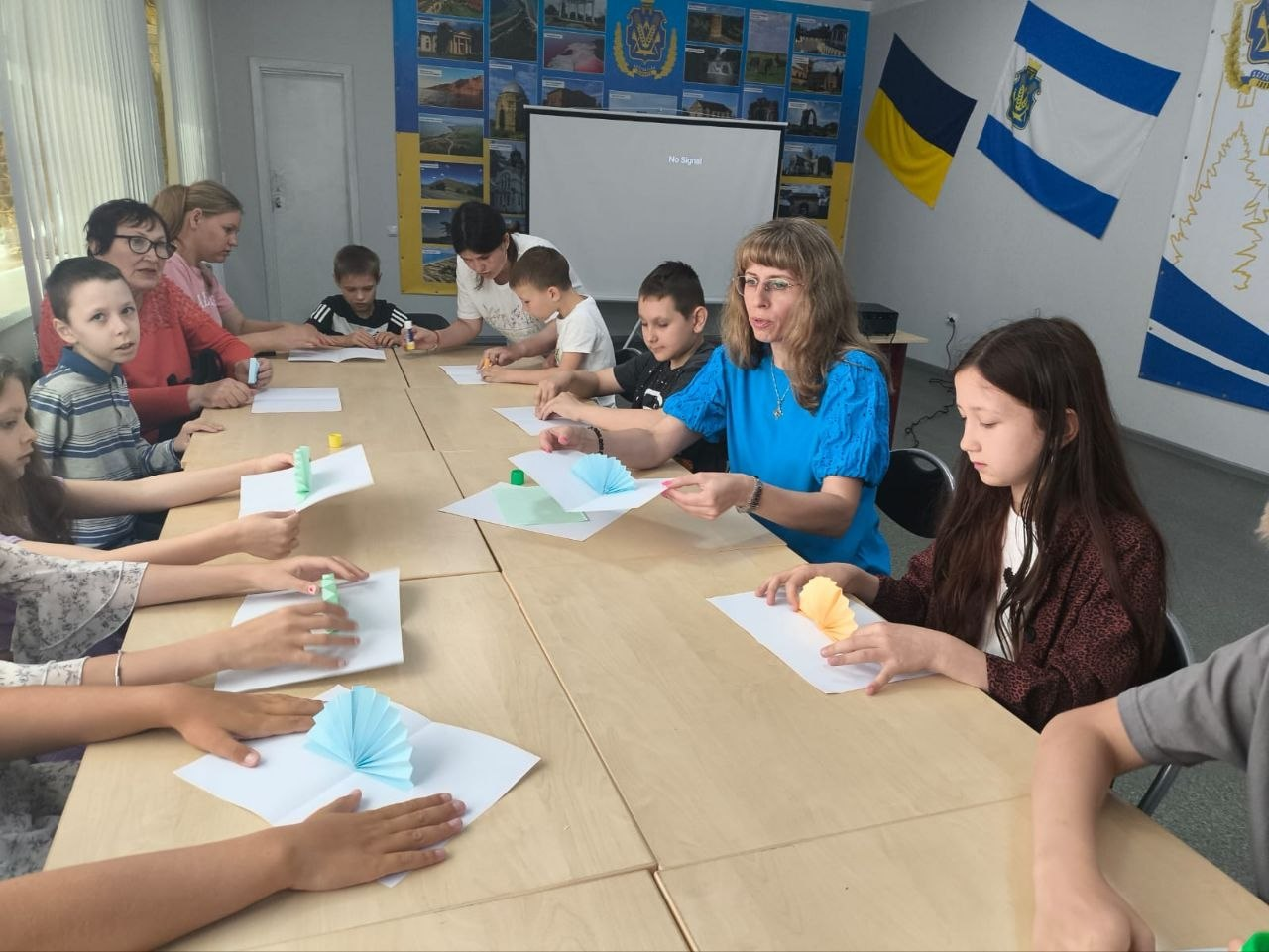 Група дітей та дорослих сидить за довгим столом у світлій кімнаті та займається творчістю. Жінка у блакитній блузці та інші учасники майструють об'ємні вироби з паперу за допомогою клею. На стінах приміщення розміщені прапори України, фотоколажі та великий білий екран.