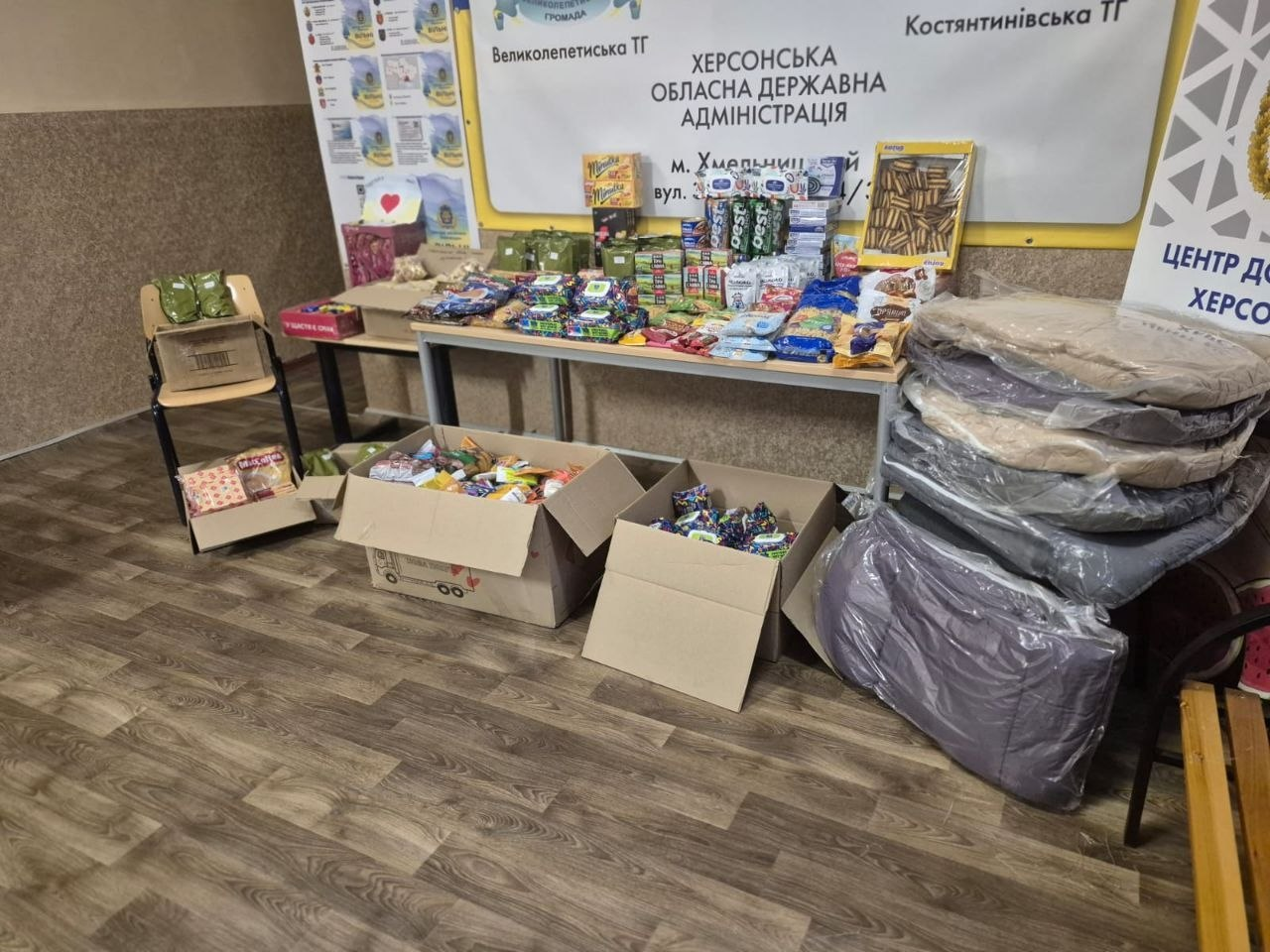 Гуманітарна допомога у приміщенні з вивісками Херсонської обласної державної адміністрації. На столах та у картонних коробках на підлозі розкладено продуктові набори, солодощі, чай та засоби гігієни. Поруч складено стоси упакованого теплого одягу або ковдр.