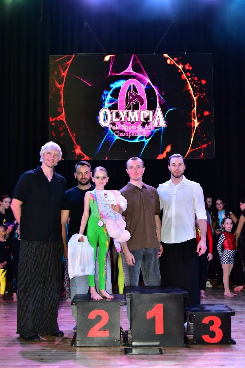 Церемонія нагородження на змаганнях «Olympia Pole Sport & Art Championship». На п'єдесталі пошани стоїть юна спортсменка в зеленому комбінезоні з дипломом, медаллю та м'якою іграшкою. Поруч із нею стоять четверо чоловіків на тлі сцени з великим екраном.